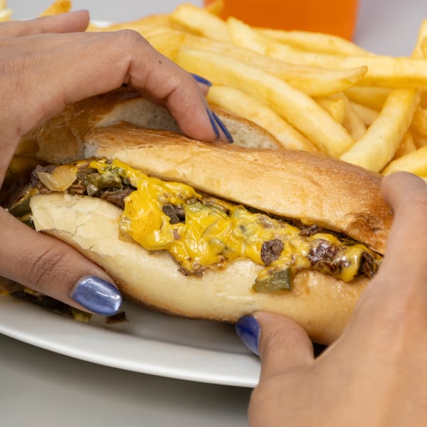 Hands holding a Seaport Diner cheesesteak sandwich with melted cheese, onions, and fries.