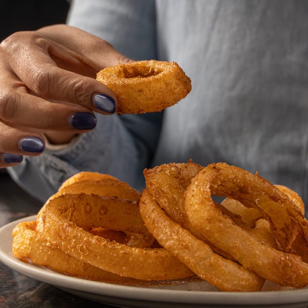 A hand with dark blue nail polish picks up a golden-brown onion ring from a white plate.